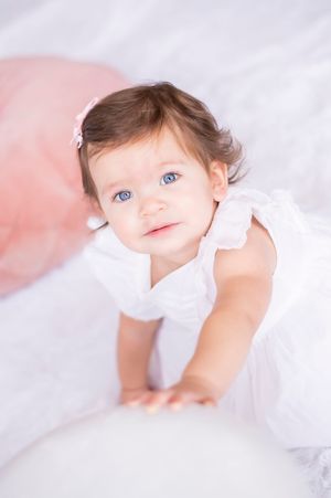 Adorable baby girl with blue eyes wearing a white dress with ruffles, looking up curiously.