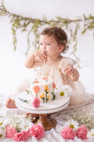 Baby girl in a floral dress smashing her first birthday cake decorated with colorful flowers.