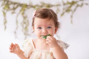 Close-up of a blue-eyed baby girl in a floral dress eating cake at her first birthday session.