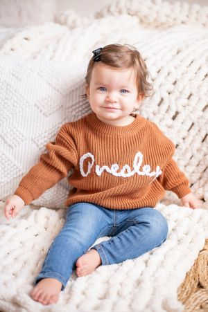 Cute toddler girl in an orange knit sweater and jeans sitting on a chunky knit blanket smiling.