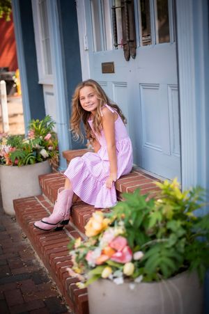 Young girl in pink gingham dress sitting on brick steps beside potted flowers near a blue front door.