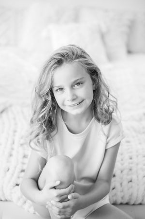 Black and white portrait of a smiling preteen girl with curly blonde hair hugging her knees.