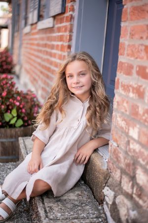 Pretty young girl with wavy blonde hair in a beige dress sitting by a brick wall outdoors.