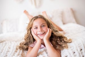 Smiling young girl with long curly blonde hair lying on a cozy chunky knit blanket on a bed.