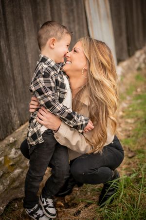 Mother crouching down hugging her young son near a weathered wooden fence outdoors.