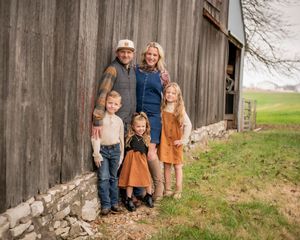 Family of five stands together beside a rustic wooden barn in the countryside.