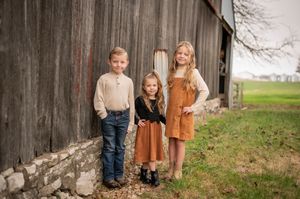 Three siblings pose against a weathered wooden barn wall in autumn outfits.