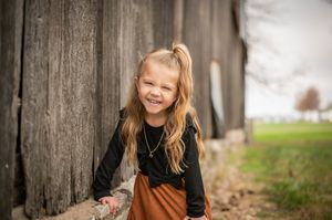 Smiling young girl with blonde ponytail leaning against rustic wooden barn wall outdoors.