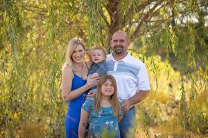 Smiling family of four posing outdoors under a weeping willow tree in golden natural light.