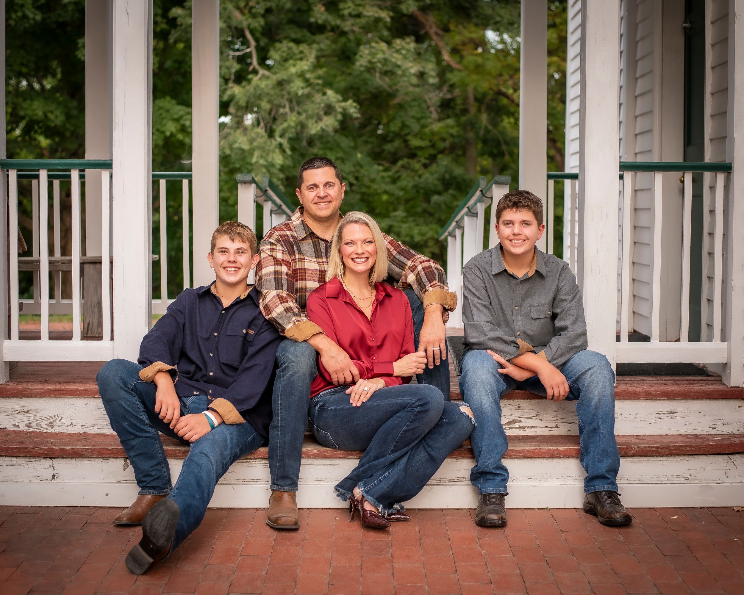 Family of four smiles together on porch steps surrounded by lush green trees.
