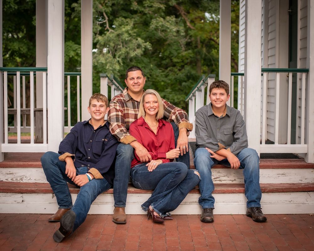 Family of four smiles together on porch steps surrounded by lush green trees.