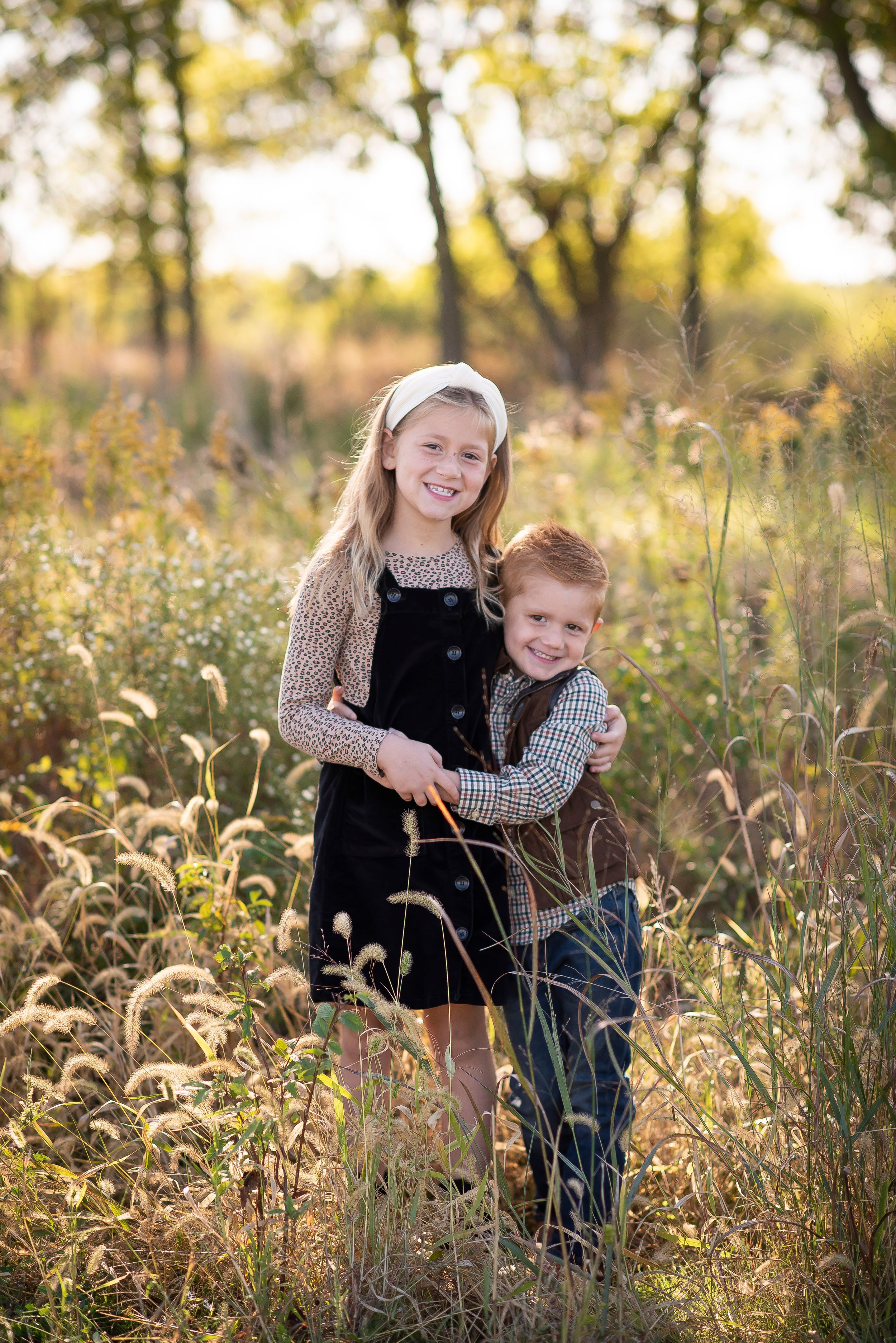 Two young siblings smiling and hugging in a sunlit field during an outdoor photo session.
