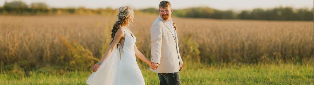 Couple holds hands while walking through golden wheat field at sunset during outdoor photo session.