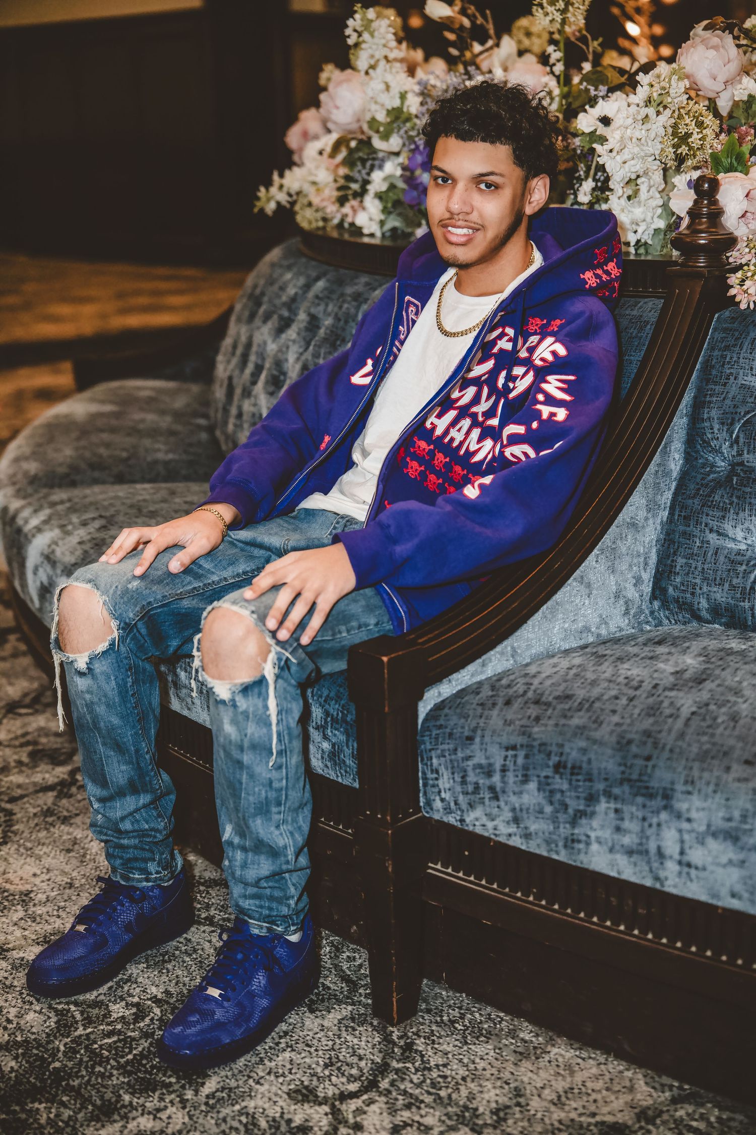 Person relaxes on velvet couch wearing blue varsity jacket and distressed jeans.
