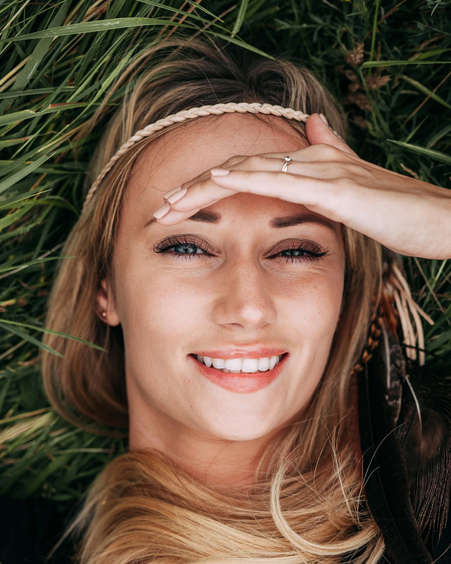 A person with blonde hair smiles with eyes closed while touching their forehead in a grassy outdoor setting.