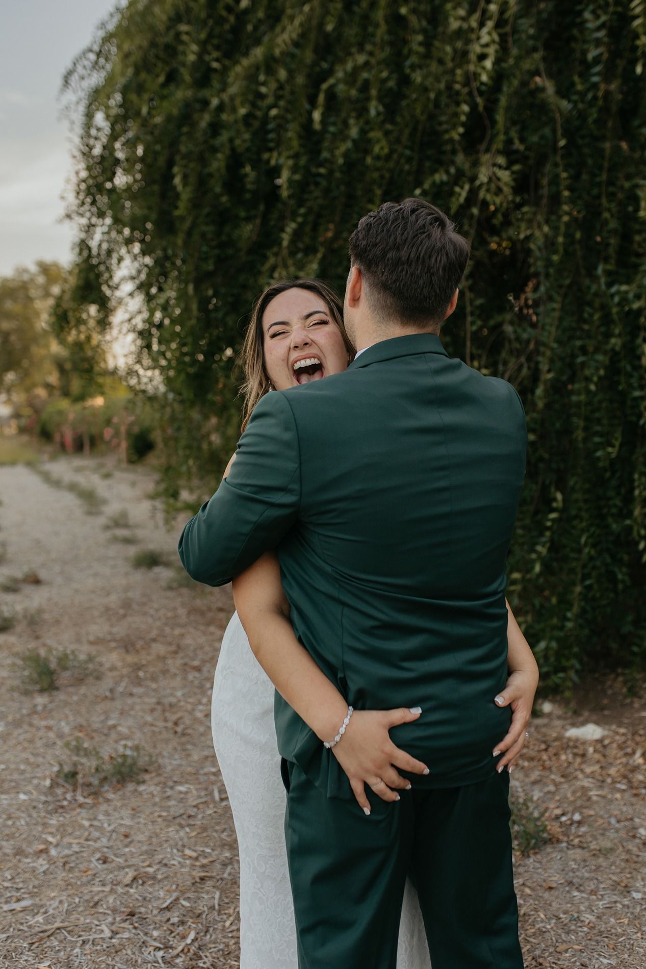 A couple embraces and laughs joyfully under a weeping willow tree during an outdoor photo session at Canopy Grove by Wedgewood Weddings.
