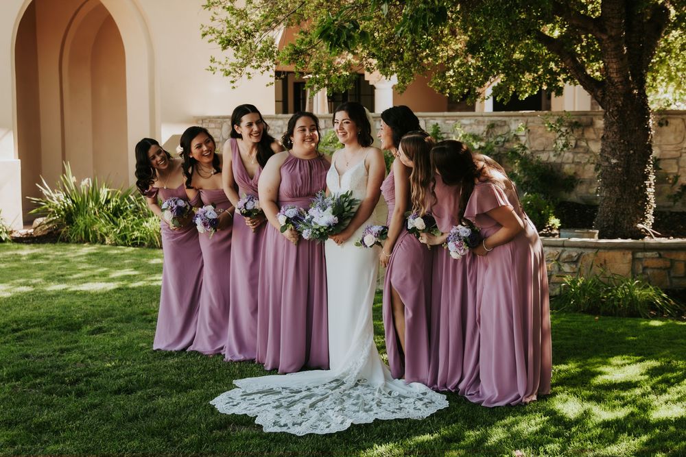 Bridal party in mauve dresses poses together on a sunny garden lawn for wedding photos.