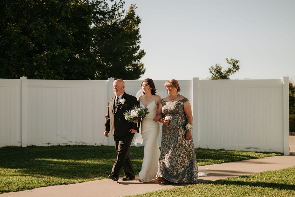 A wedding party walks down an outdoor path alongside a white fence during a golden sunset ceremony.
