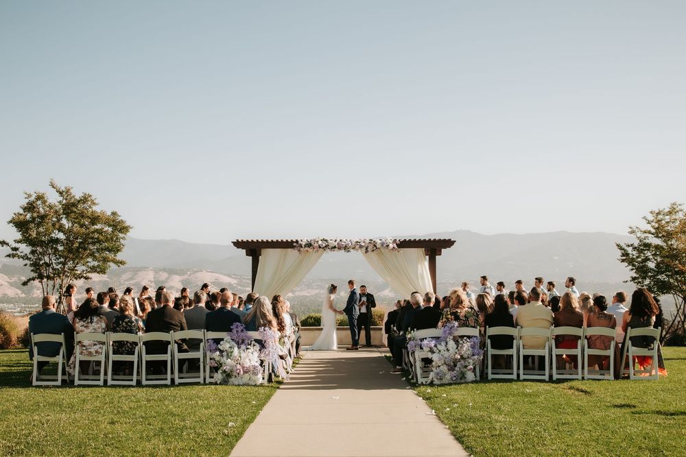 Panoramic view of wedding guests seated in white chairs on a hilltop overlooking a scenic landscape.