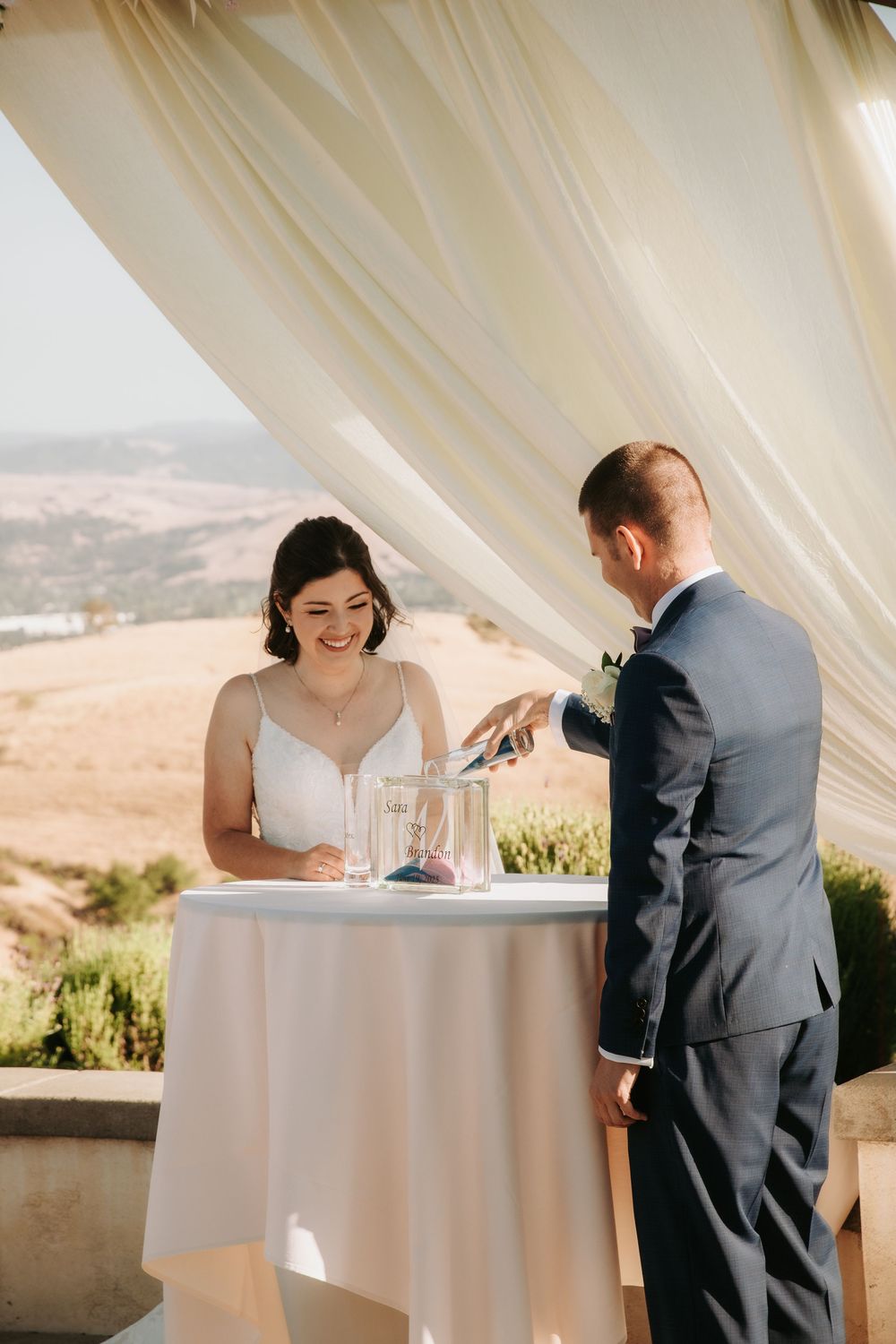 A couple shares an intimate moment at a signing table draped in white cloth with flowing curtains and scenic views.