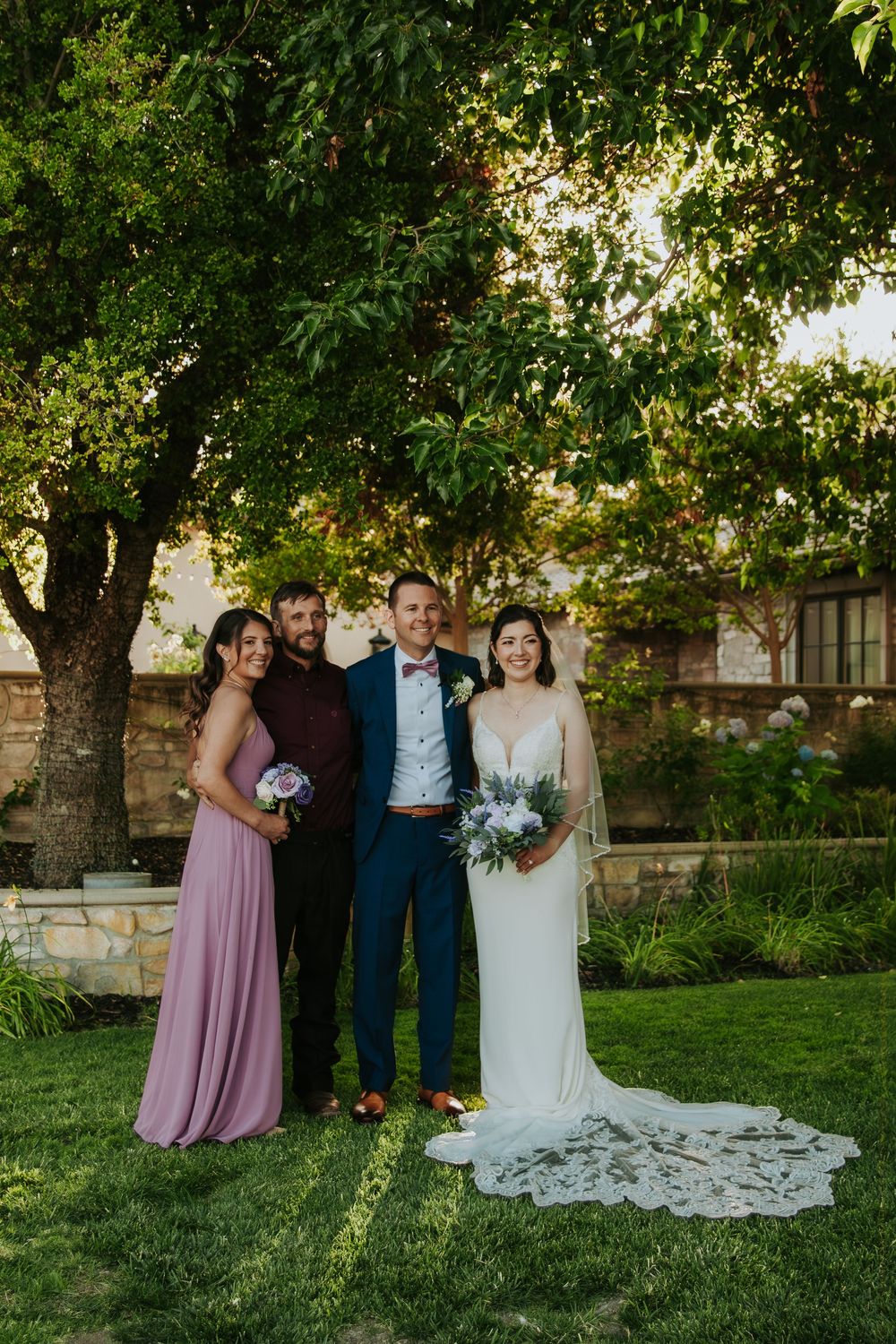 Wedding party poses for photos on a lush green lawn with trees in the background.