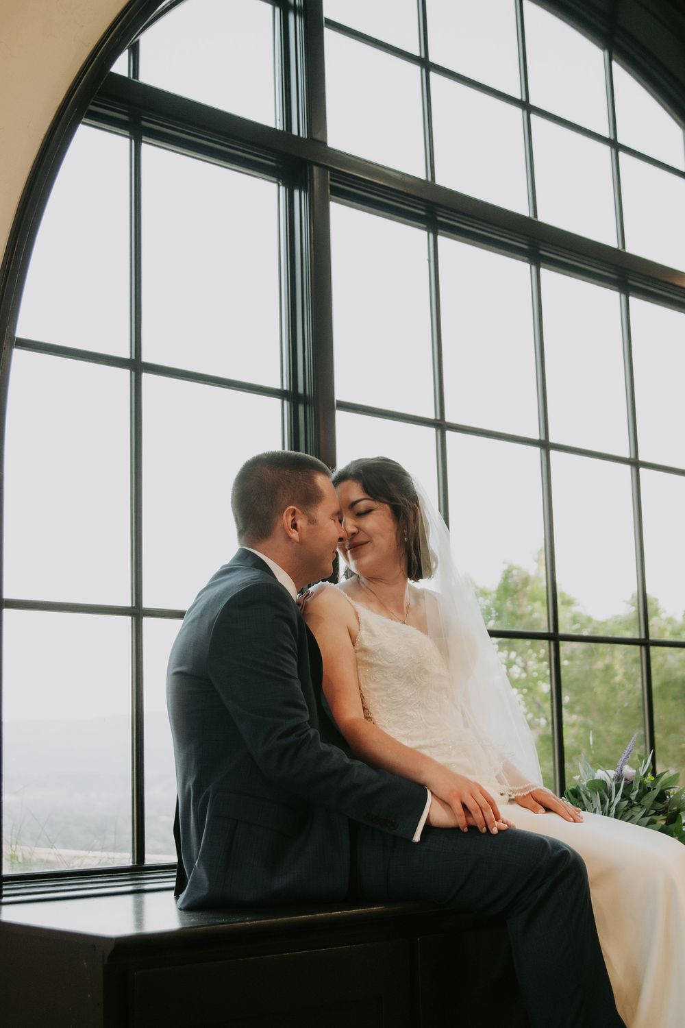 A tender wedding moment shared beside a large arched window overlooking scenic views.
