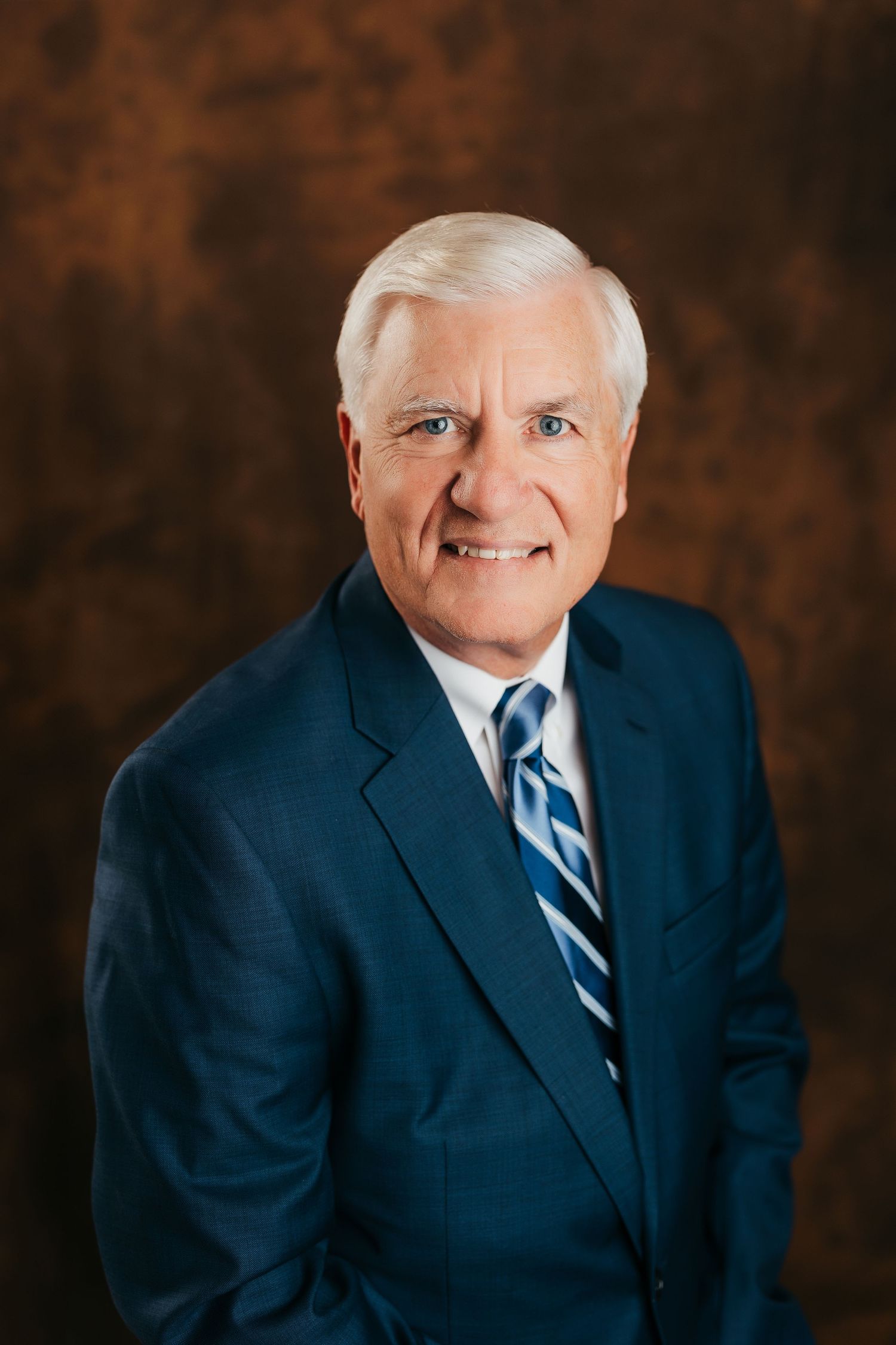 Professional headshot of a person in a navy suit and striped tie against a dark brown background.