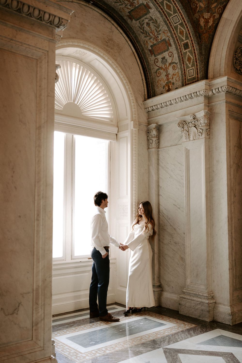 Couple holds hands facing each other by arched window in marble Library of Congress corridor.
