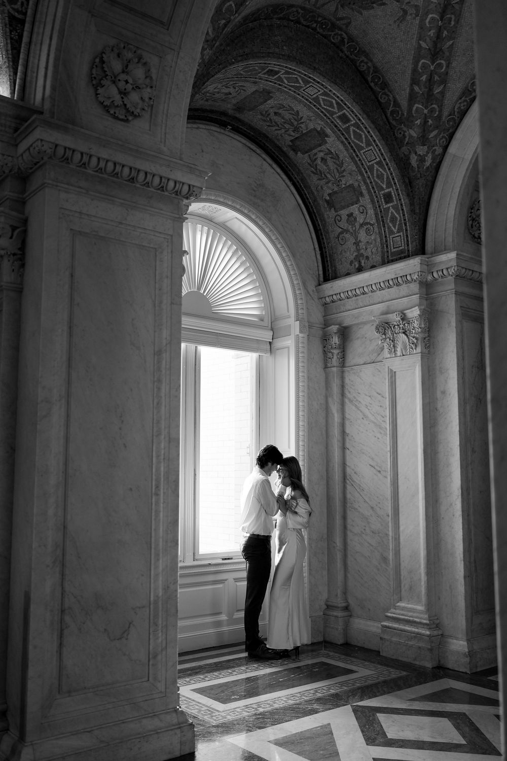 Black and white photo of couple embracing by arched window in ornate marble building.