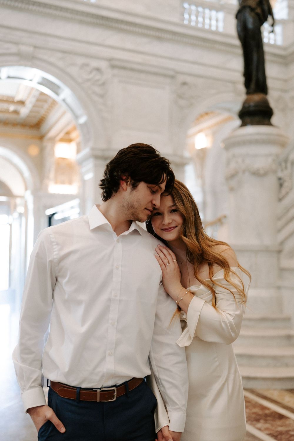 Couple embraces warmly inside grand marble hall with classical architecture and decorative statues.