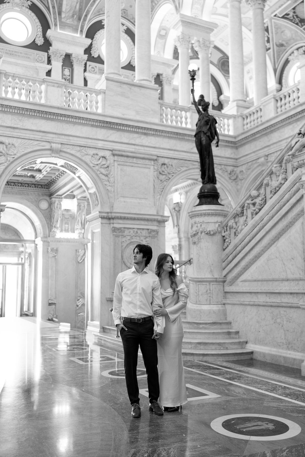 Couple stands together in black and white photo inside ornate marble hall with grand staircase and classical statue.