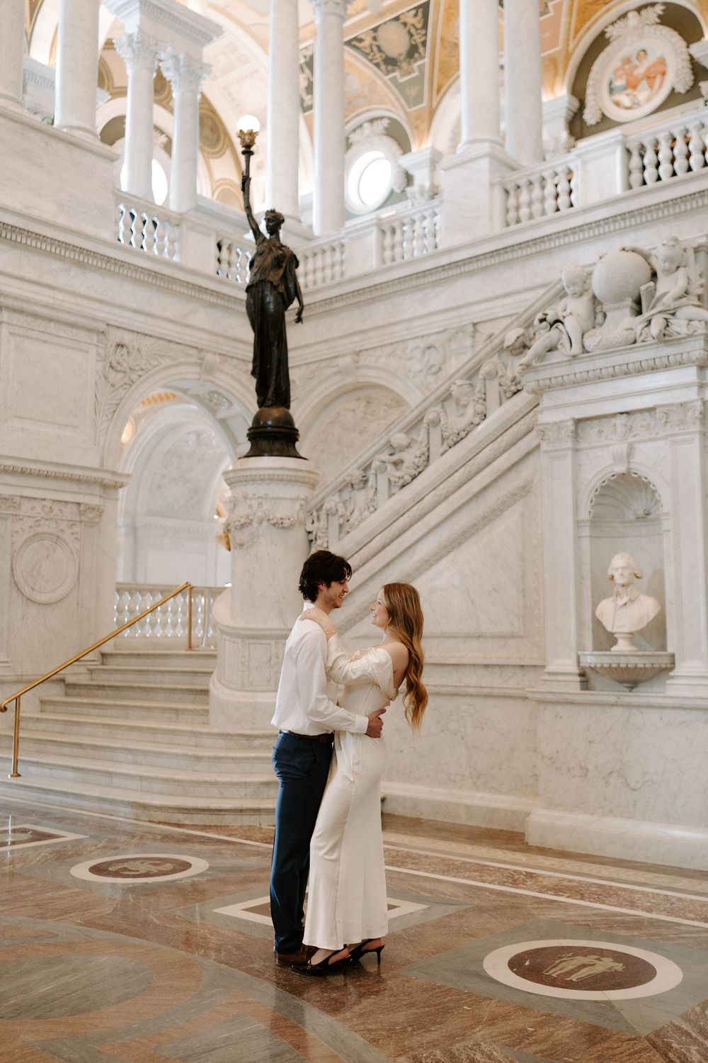 Couple embraces near a bronze statue on the ornate marble staircase of the Library of Congress.