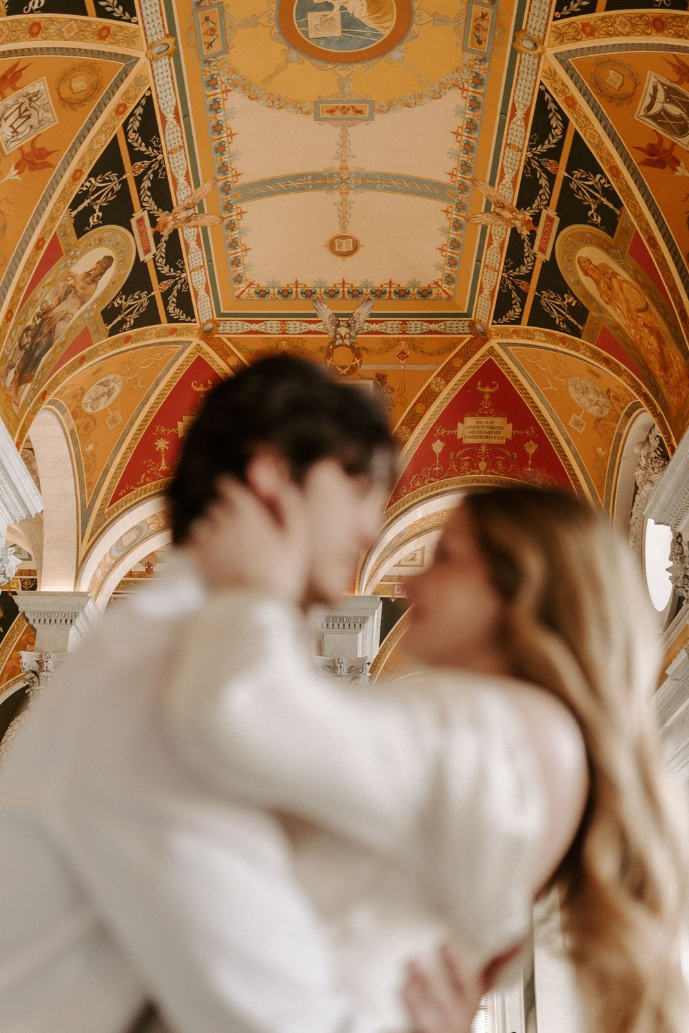 Couple shares a kiss beneath stunning painted vaulted ceiling with colorful frescoes in historic building.