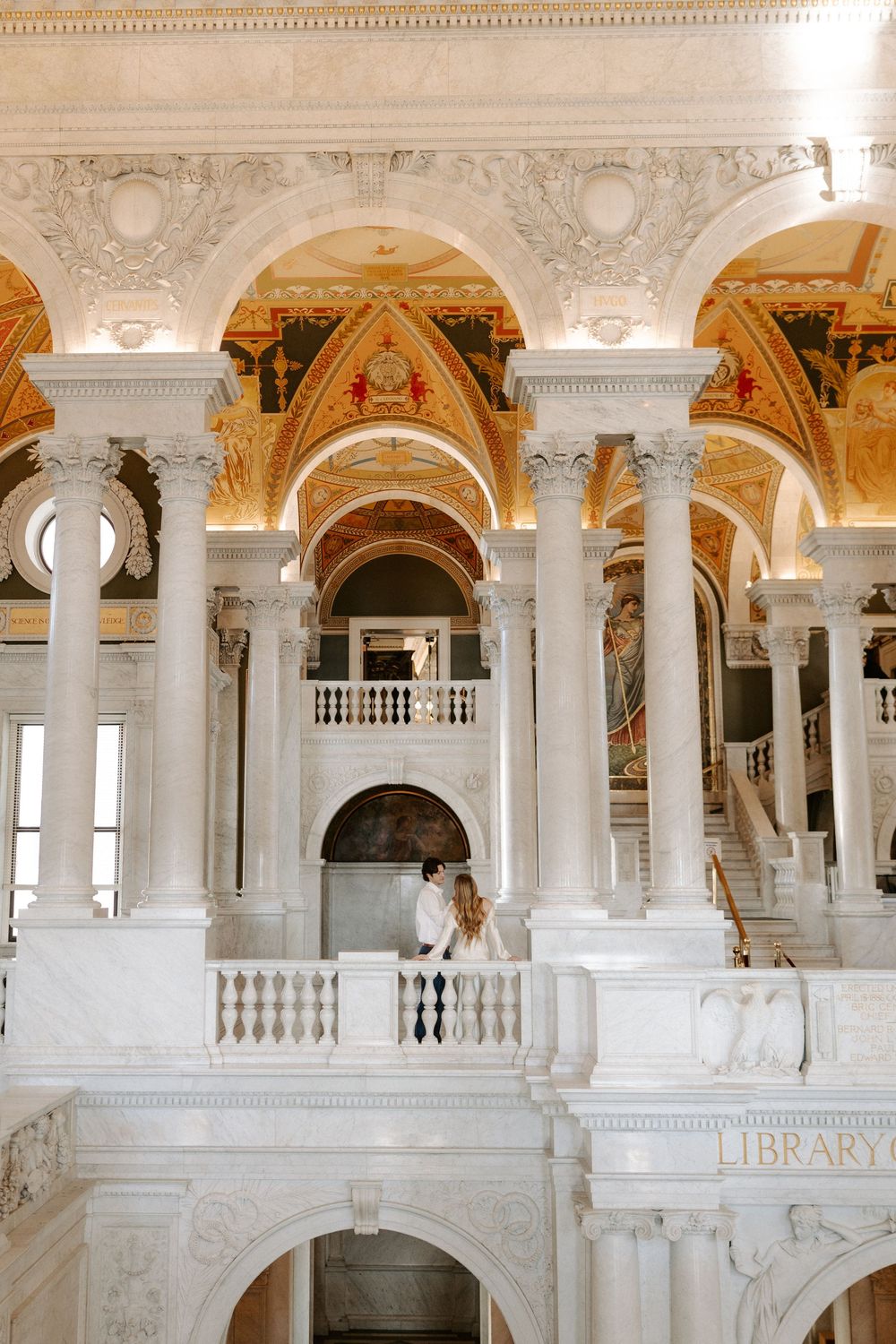 Couple embraces on marble balcony inside grand library with soaring arches, columns, and gilded ceiling.