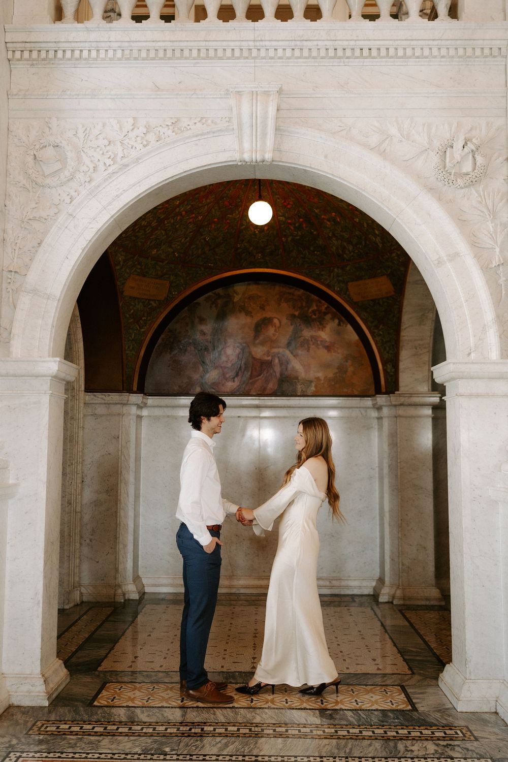 Couple holds hands facing each other beneath ornate stone arch with classical mural in elegant marble hall.