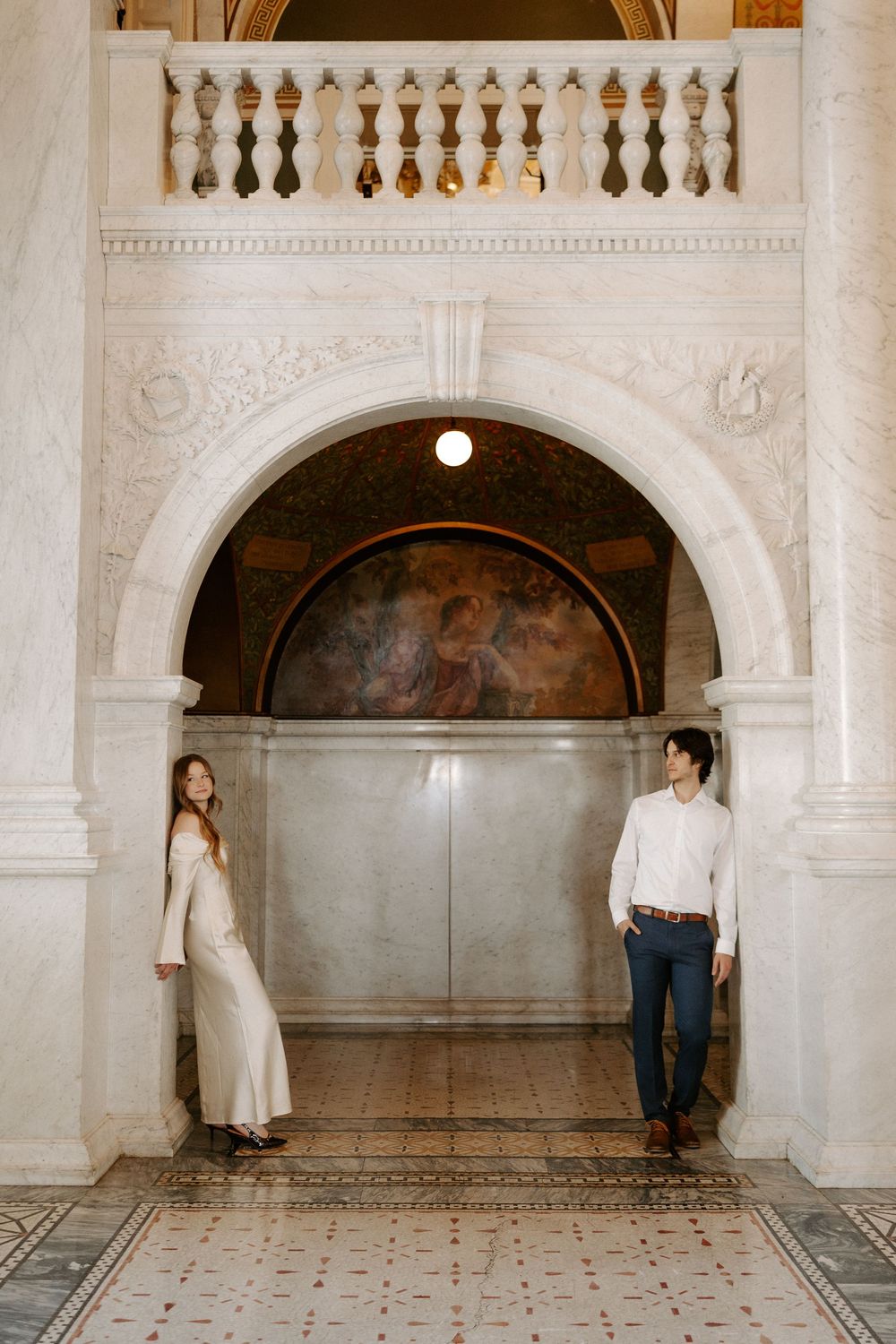 Couple stands apart beneath decorative arch with Renaissance-style mural in a grand marble interior.