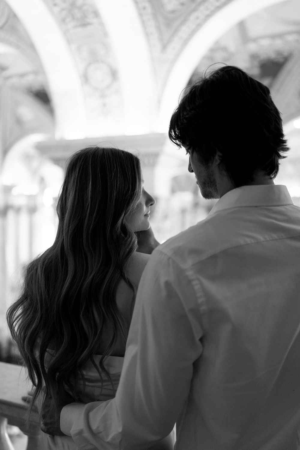 Black and white portrait of couple embracing, woman with long flowing hair in elegant arched hallway.