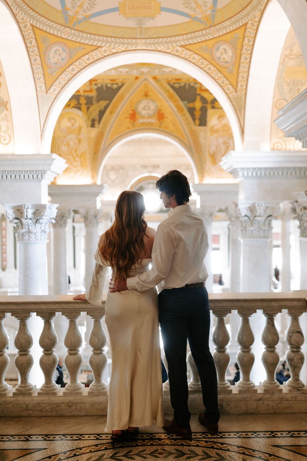 Couple stands arm in arm at marble balustrade beneath gilded arched ceiling in grand classical interior.