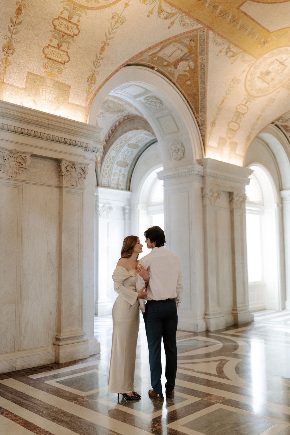 Couple embraces in ornate marble corridor with gilded arched ceiling and decorative mosaic tile floor.