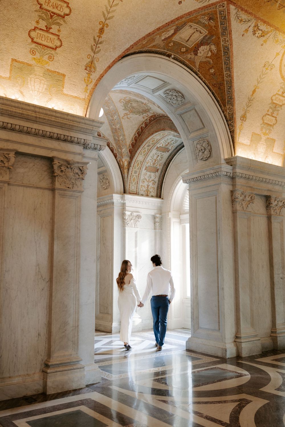 Couple walks hand in hand through ornate marble archway with gilded ceiling in historic library.