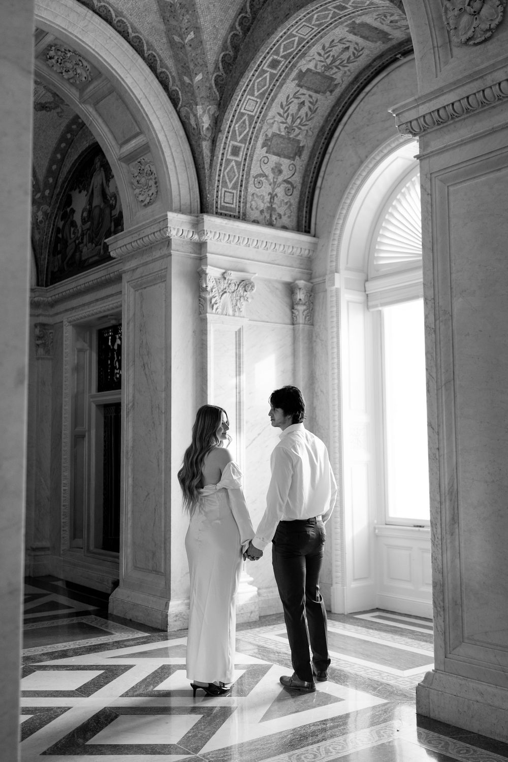 Couple holds hands in black and white photo inside ornate marble hallway with arched windows and carved ceiling.