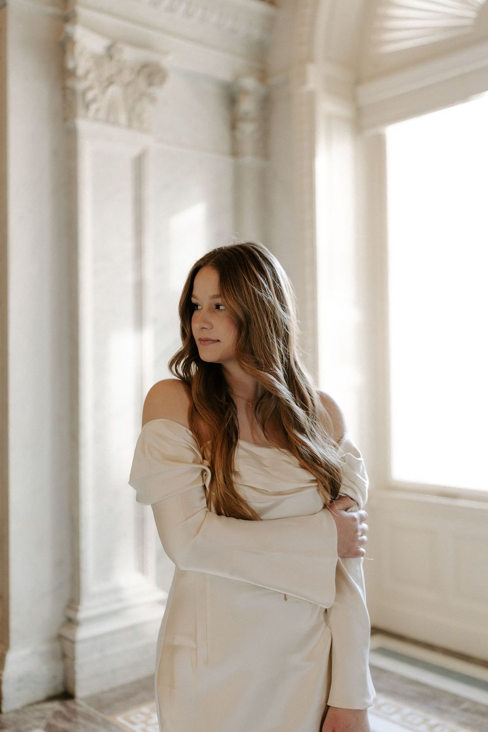 Young woman in off-shoulder white dress poses gracefully among elegant marble columns in a classical architectural setting.
