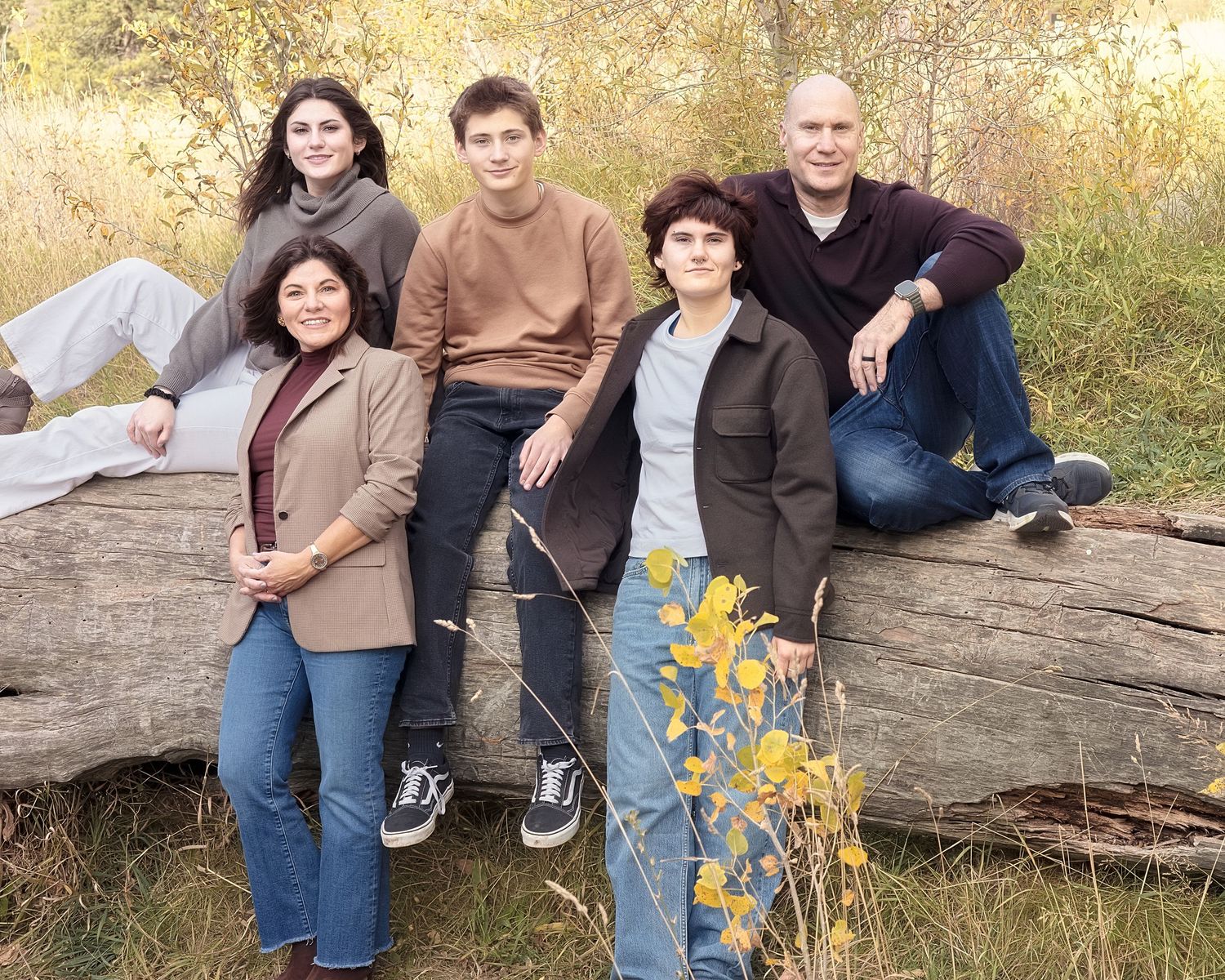 A group sits together on a fallen log in an autumn outdoor setting with golden foliage in the background.
