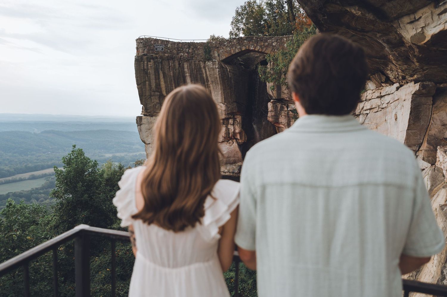 A romantic wedding couple shares an intimate moment on a mountain cliff overlooking a scenic valley landscape.