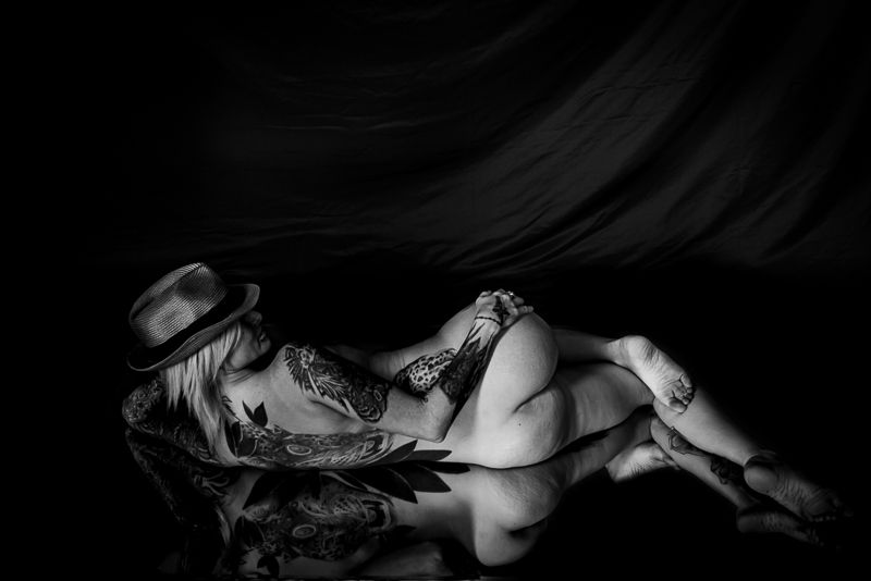 Black and white artistic boudoir photograph featuring a reclining figure in a hat against a dark background.