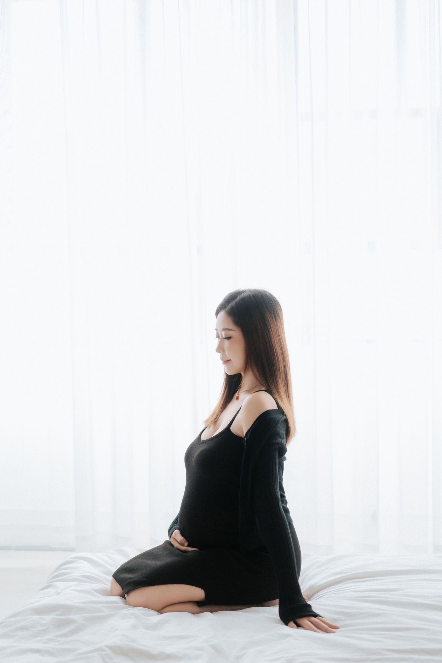 A person in black attire sits on a white bed near sheer curtains in a bright, minimalist setting.