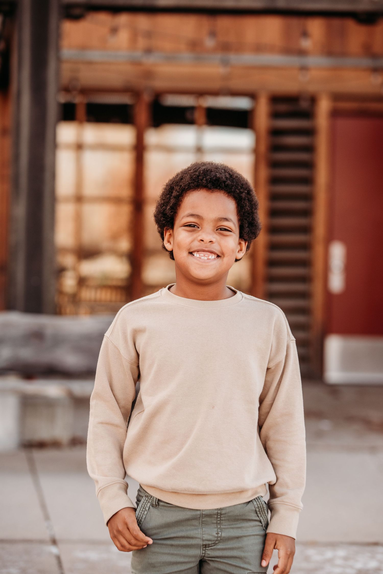 A young person in a beige sweater smiling brightly in front of a rustic wooden building.