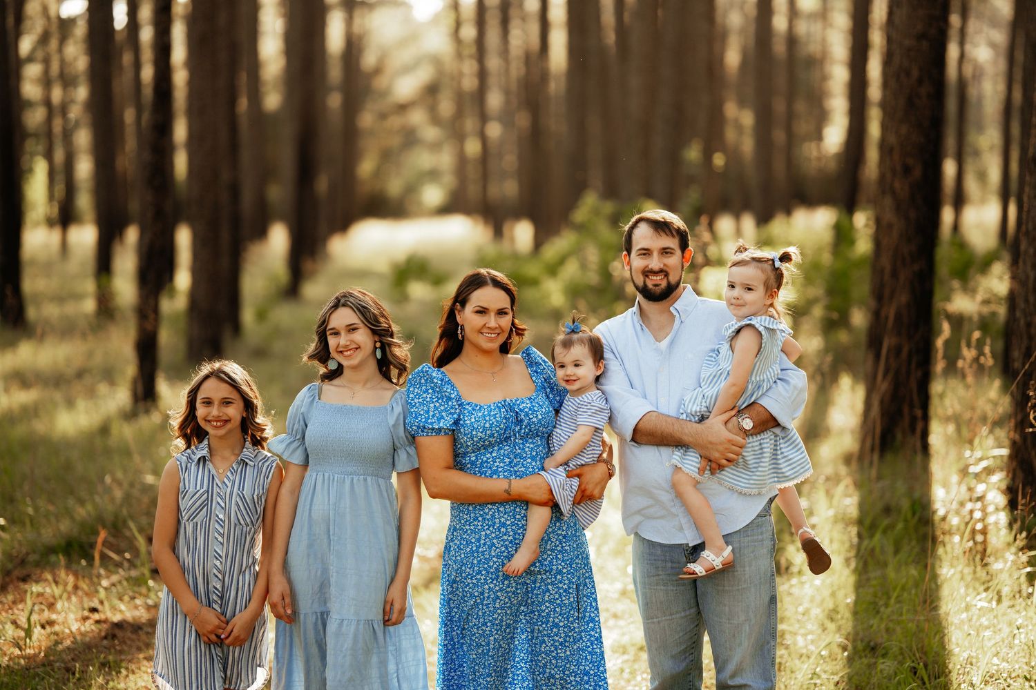 A family wearing coordinating blue and white outfits poses together in a sunny forest setting.