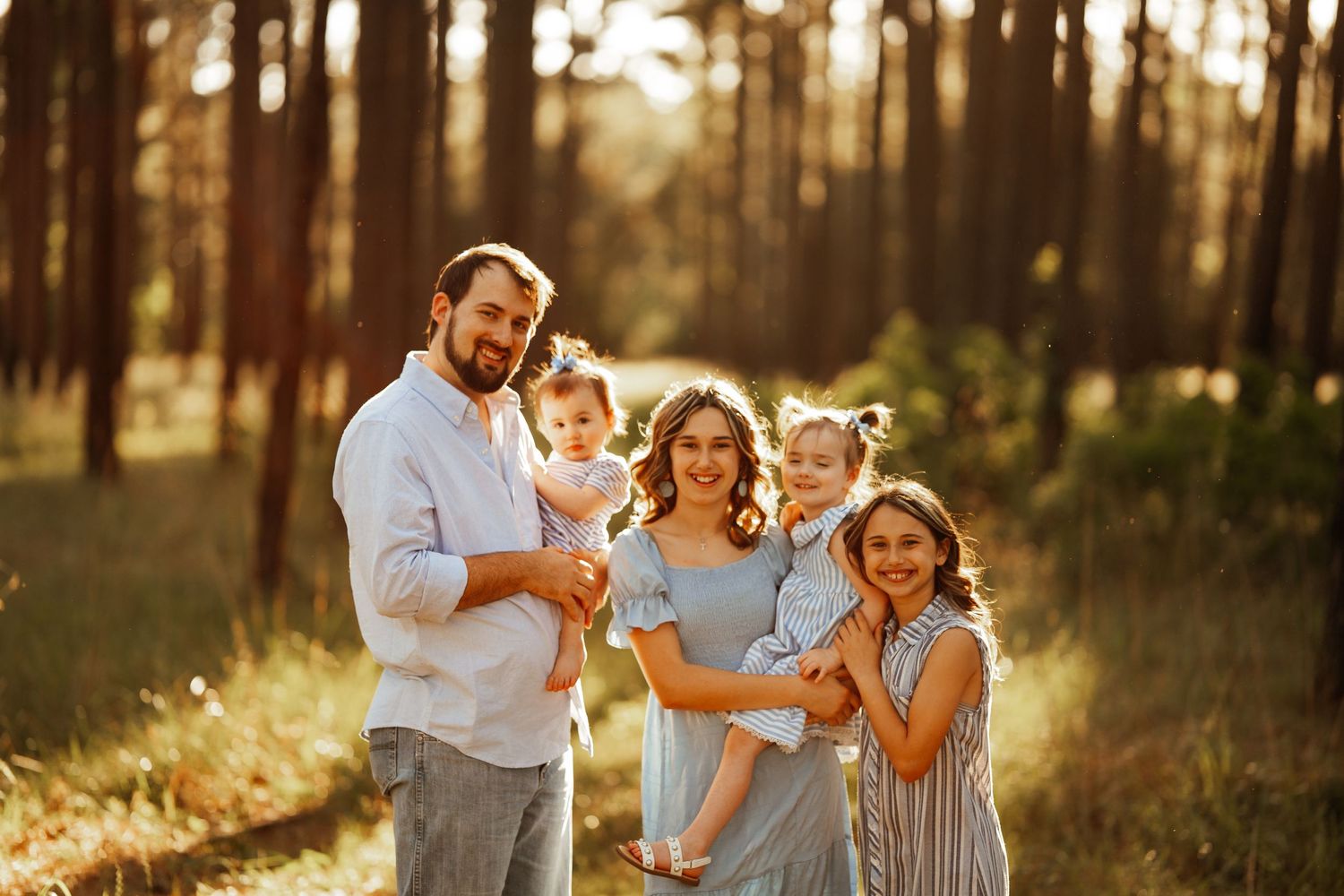 A family of four stands together in a sunlit forest during golden hour in casual attire.