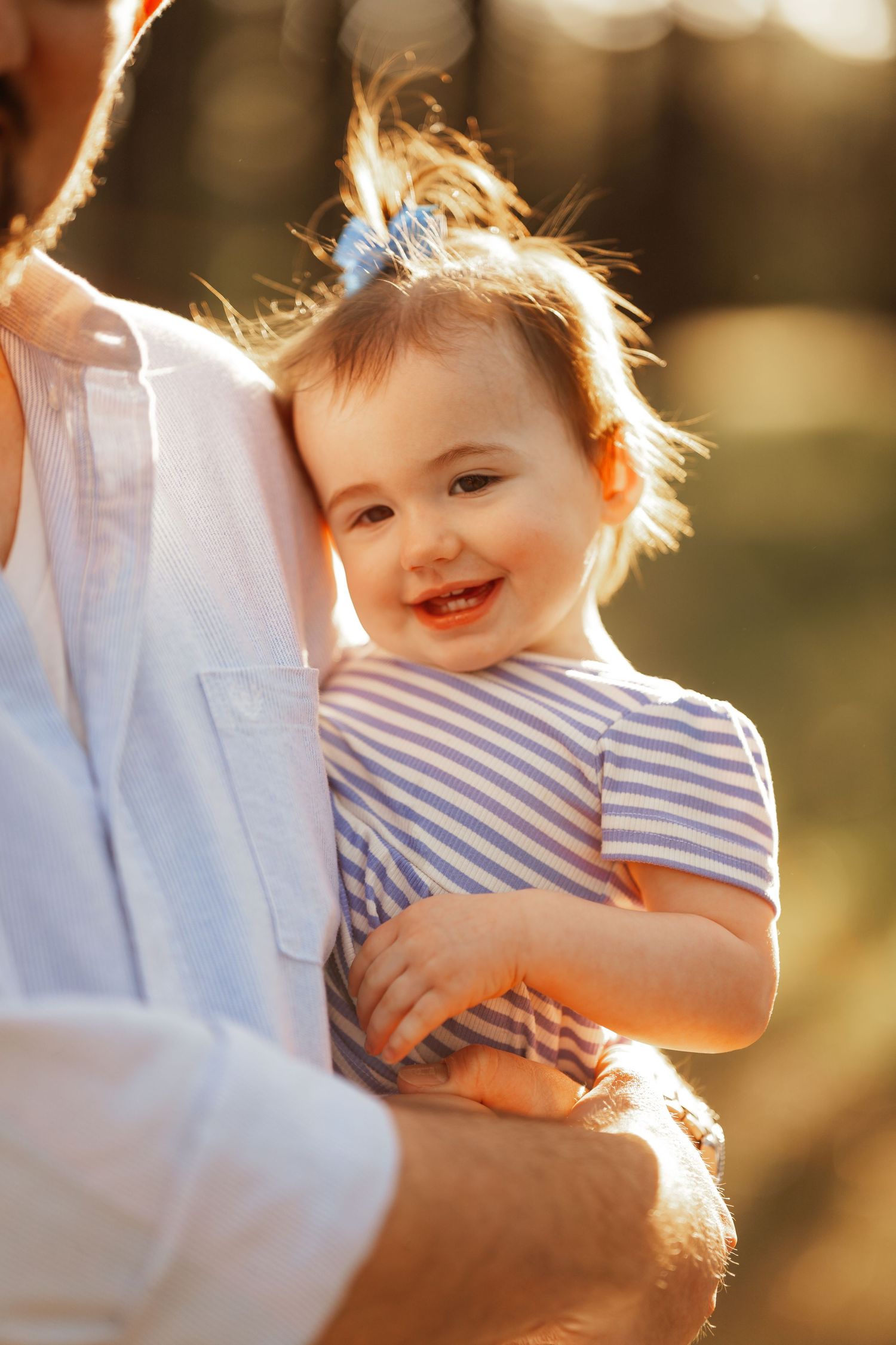 A smiling baby in a striped shirt glows in golden sunlight while being held by someone in white clothing.